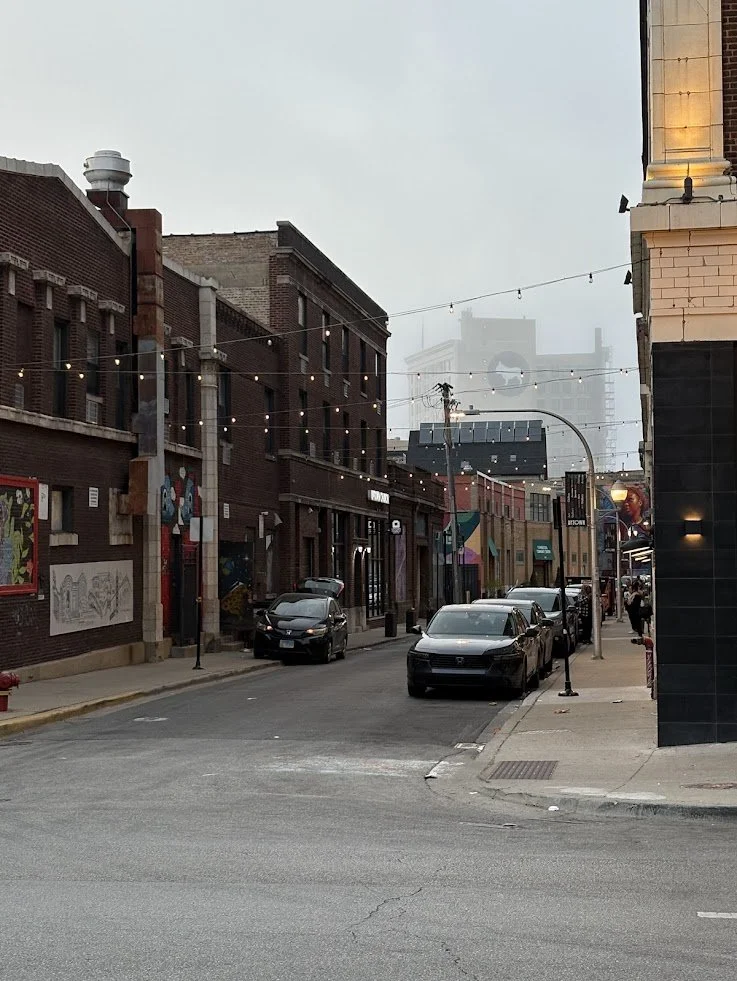 City street with brick buildings, parked cars, street lamps, string lights, and foggy weather.