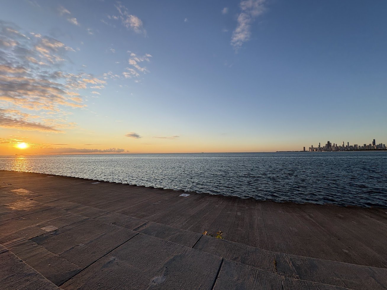 Sunset over a body of water with a city skyline in the distance and a concrete shoreline in the foreground.