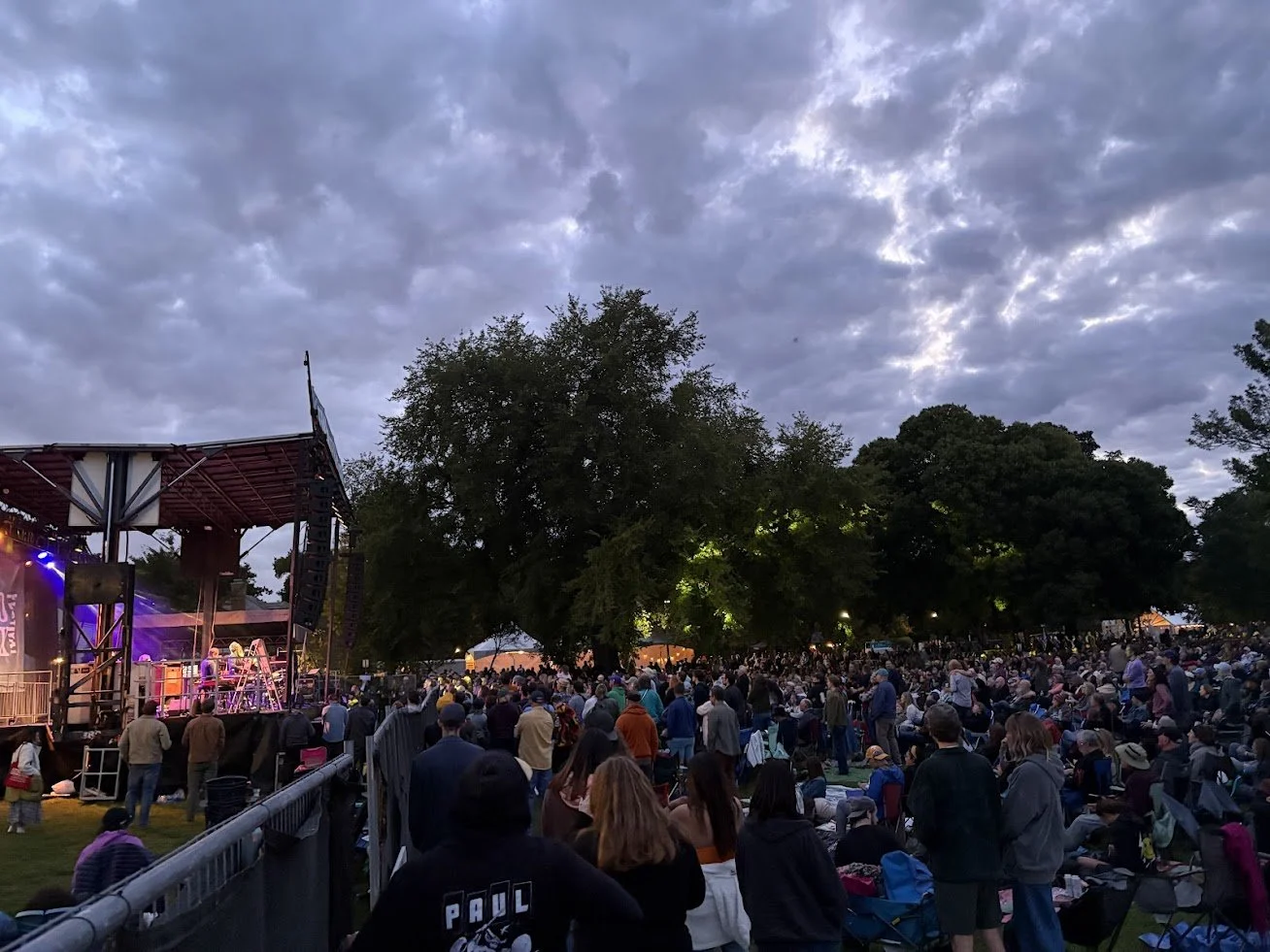 Large outdoor concert with many people gathered around a stage under a cloudy evening sky, trees surrounding the area.