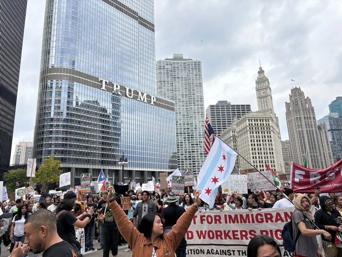 Protesters gather on a city street holding signs and flags in front of tall buildings, including the Trump Tower in Chicago, during daytime.