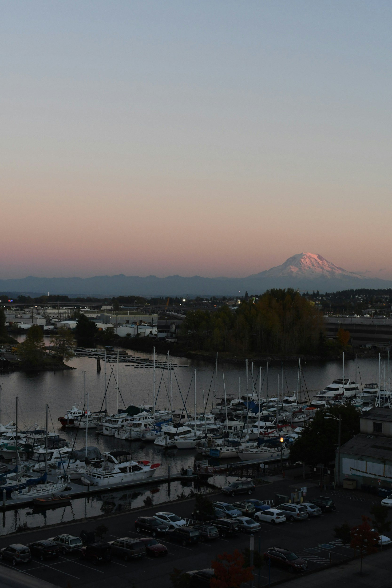 A harbor with multiple docked boats and yachts, a parking lot in the foreground, trees, industrial buildings in the middle ground, and snow-capped mountains in the background at sunset.