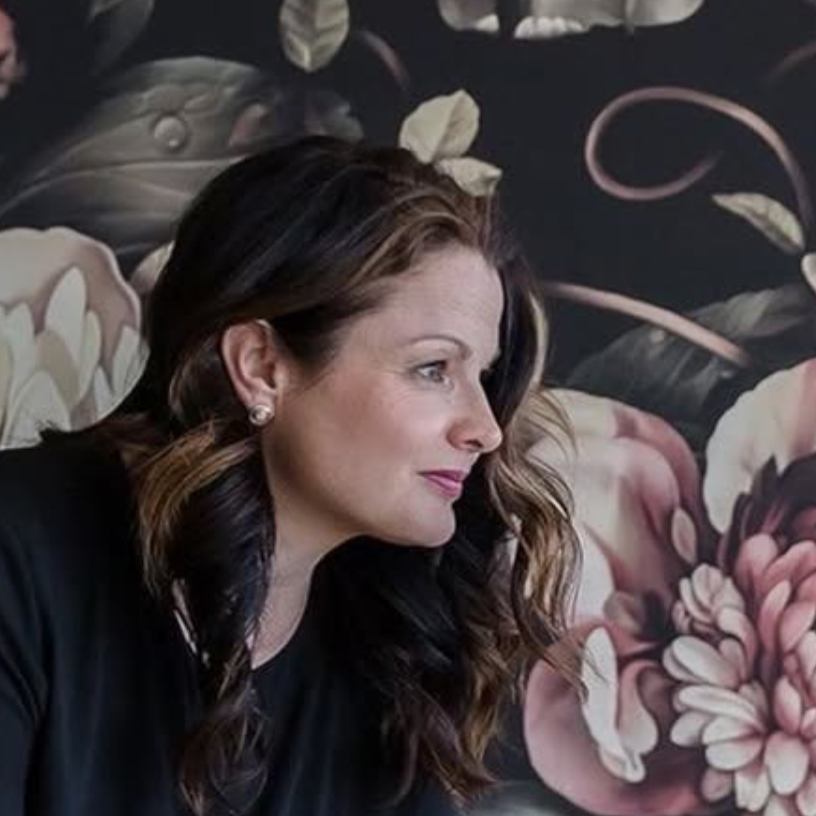 A woman with wavy brown hair and earrings, sitting against a floral patterned wall.