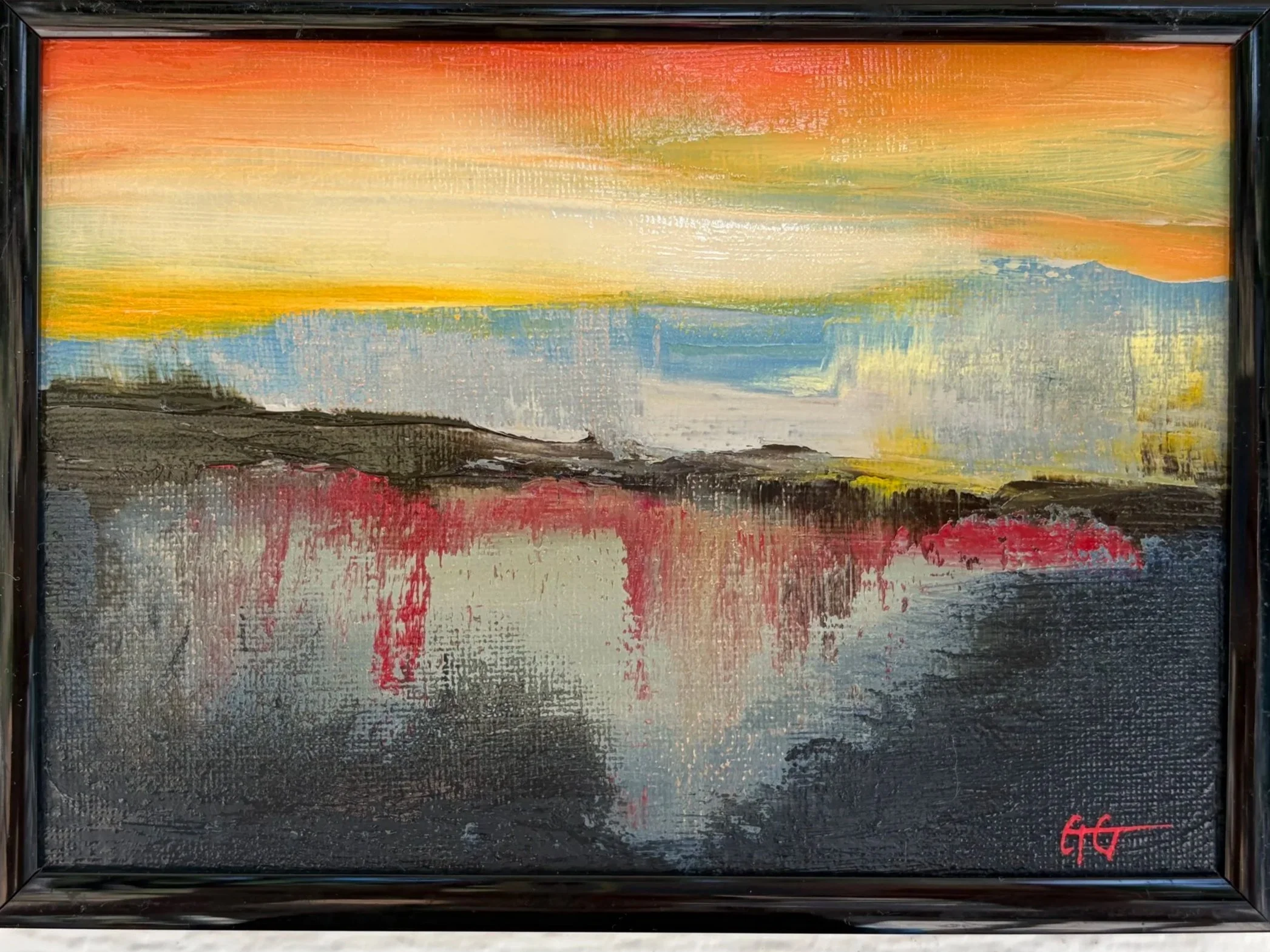 Abstract landscape painting with a sunset sky in shades of orange, yellow, and cream above a lake with reflections of red, black, and blue on the water, black frame.