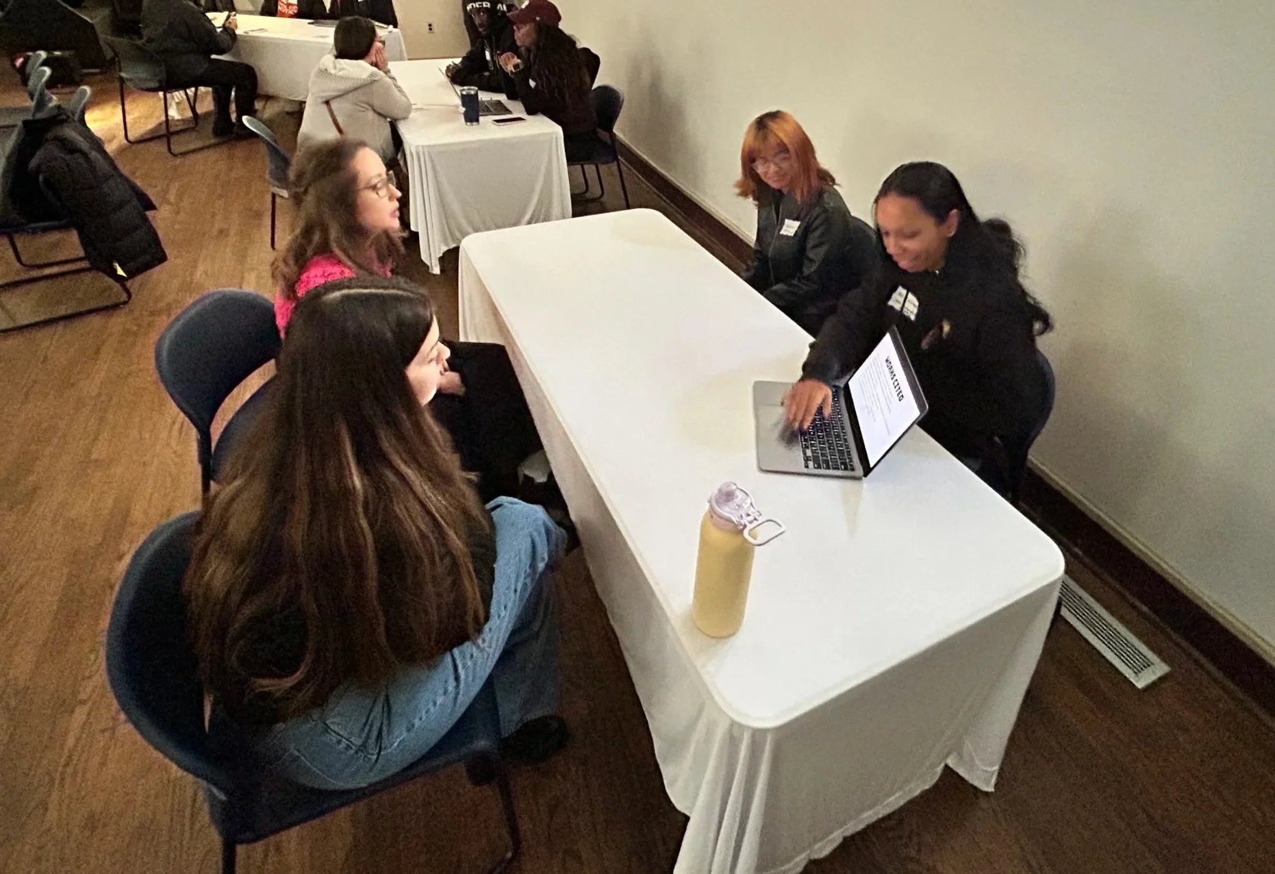 Group of women sitting at a table during a conference or meeting, with one woman using a laptop, and others listening or talking.