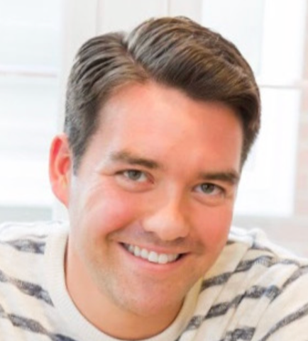 Close-up of a smiling young man with short brown hair, wearing a striped shirt, in a bright room with windows in the background.