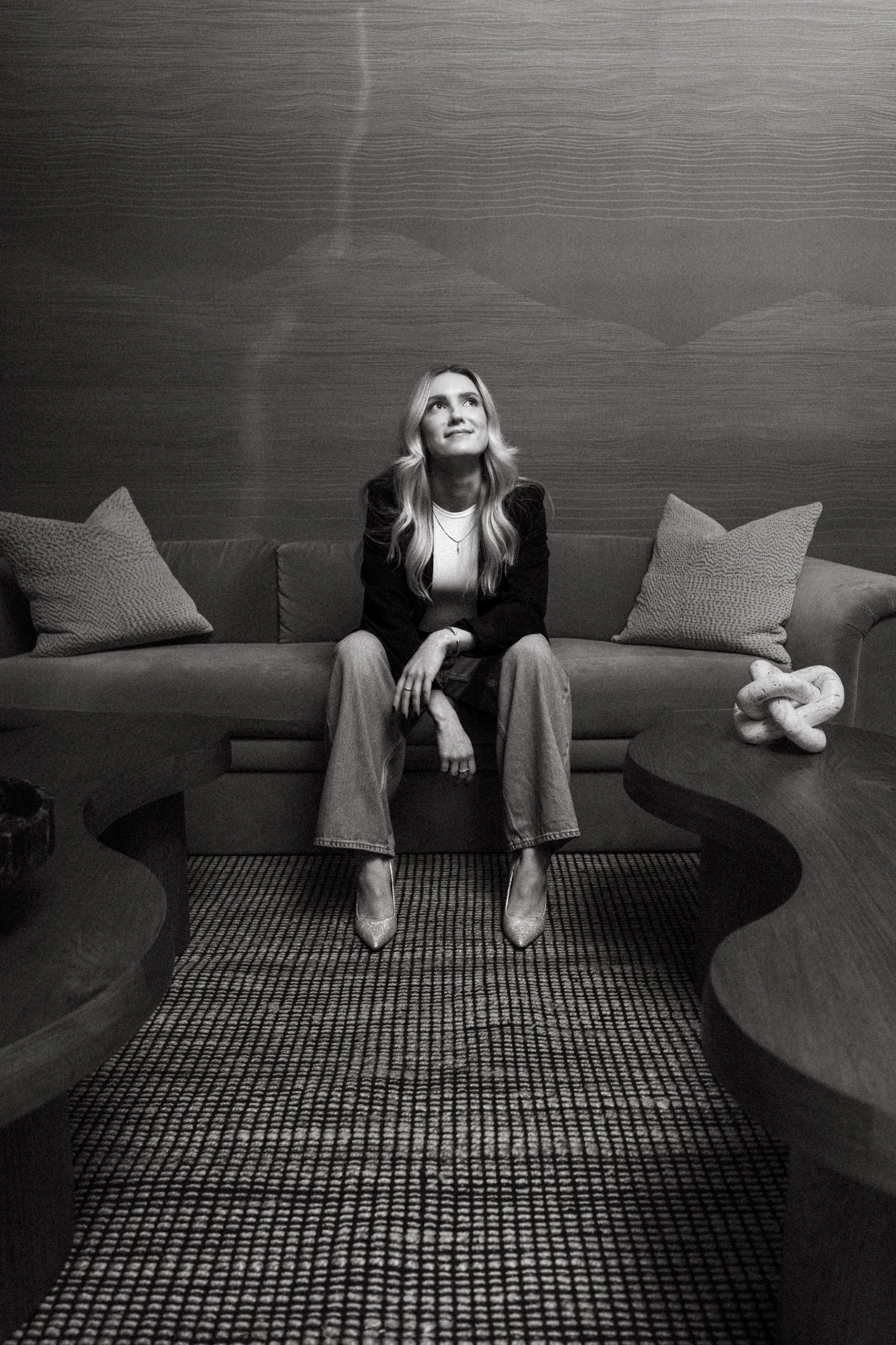 A woman sitting on a sofa in a living room, looking up with a slight smile, wearing a blazer, jeans, and heels, with pillows on the sofa and a knot-shaped sculpture on a nearby table. Brand portraiture in luxury hotel. Washington D.C.