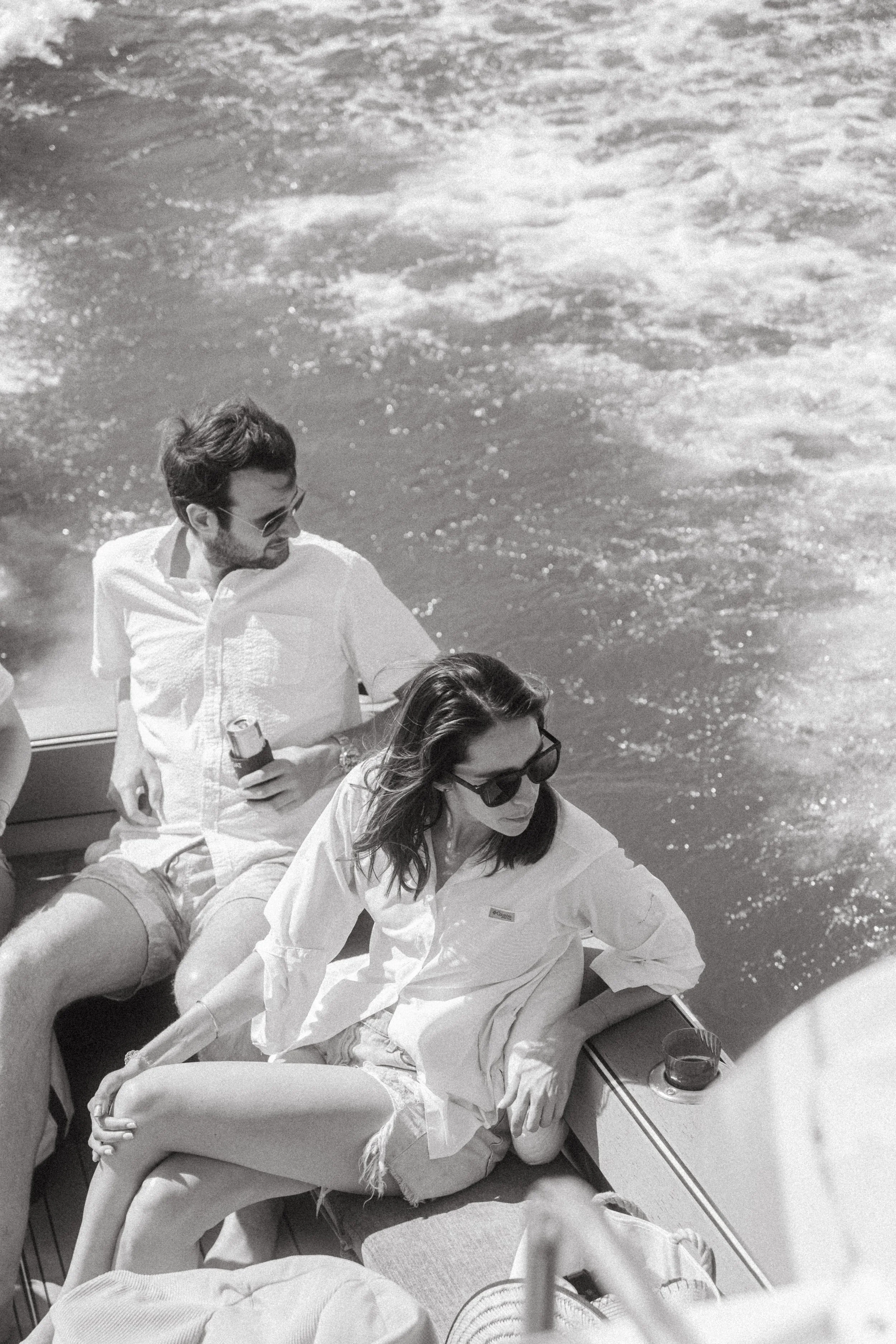 Two people on a boat, one man and one woman, both wearing sunglasses and casual clothing, sitting on the boat near water with foamy waves in the background. Yacht Cruise Washington D.C.
