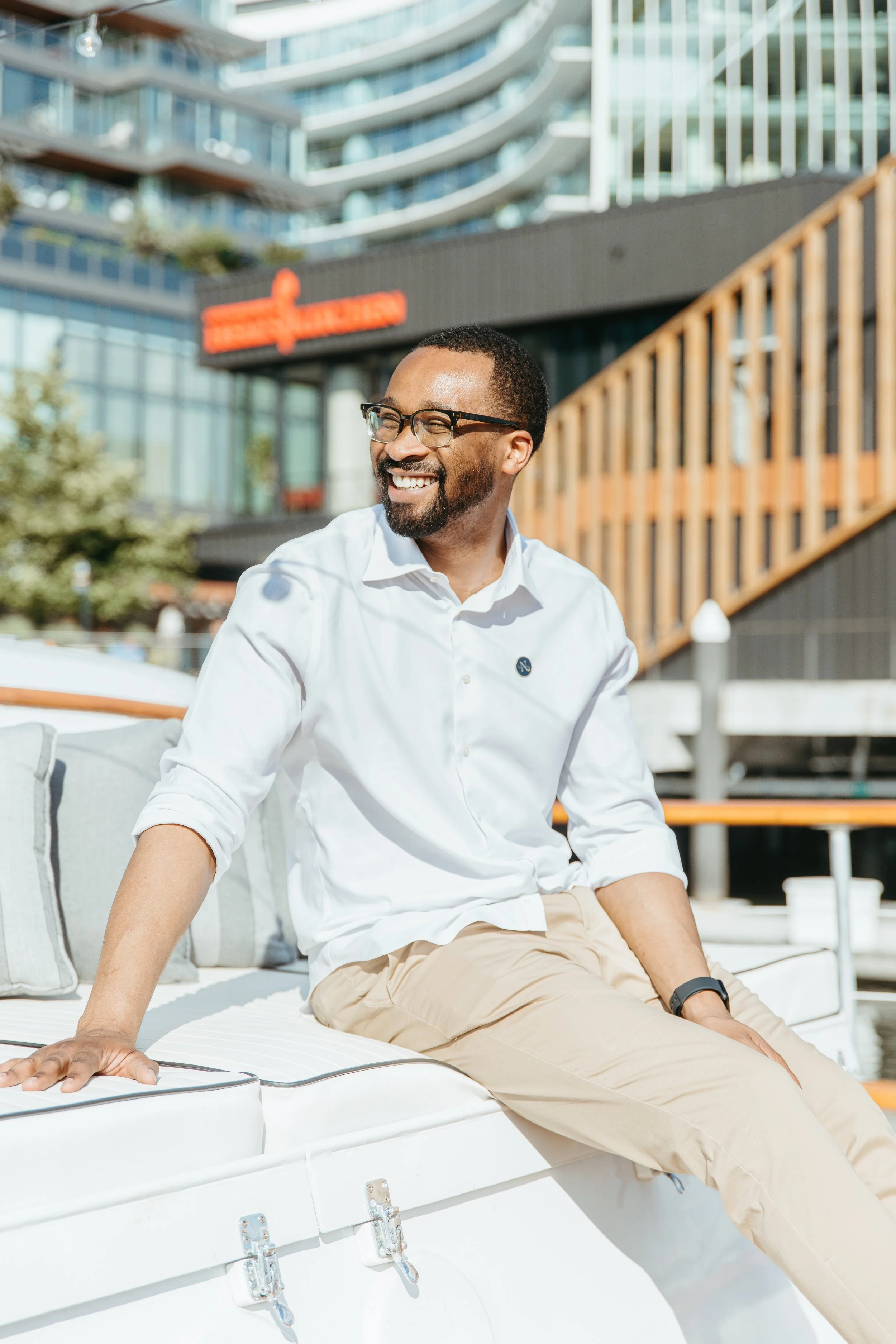 A man with glasses and a beard smiling while sitting outdoors on a boat or boat-like structure with modern buildings in the background. Yacht Cruise in Washington D.C.