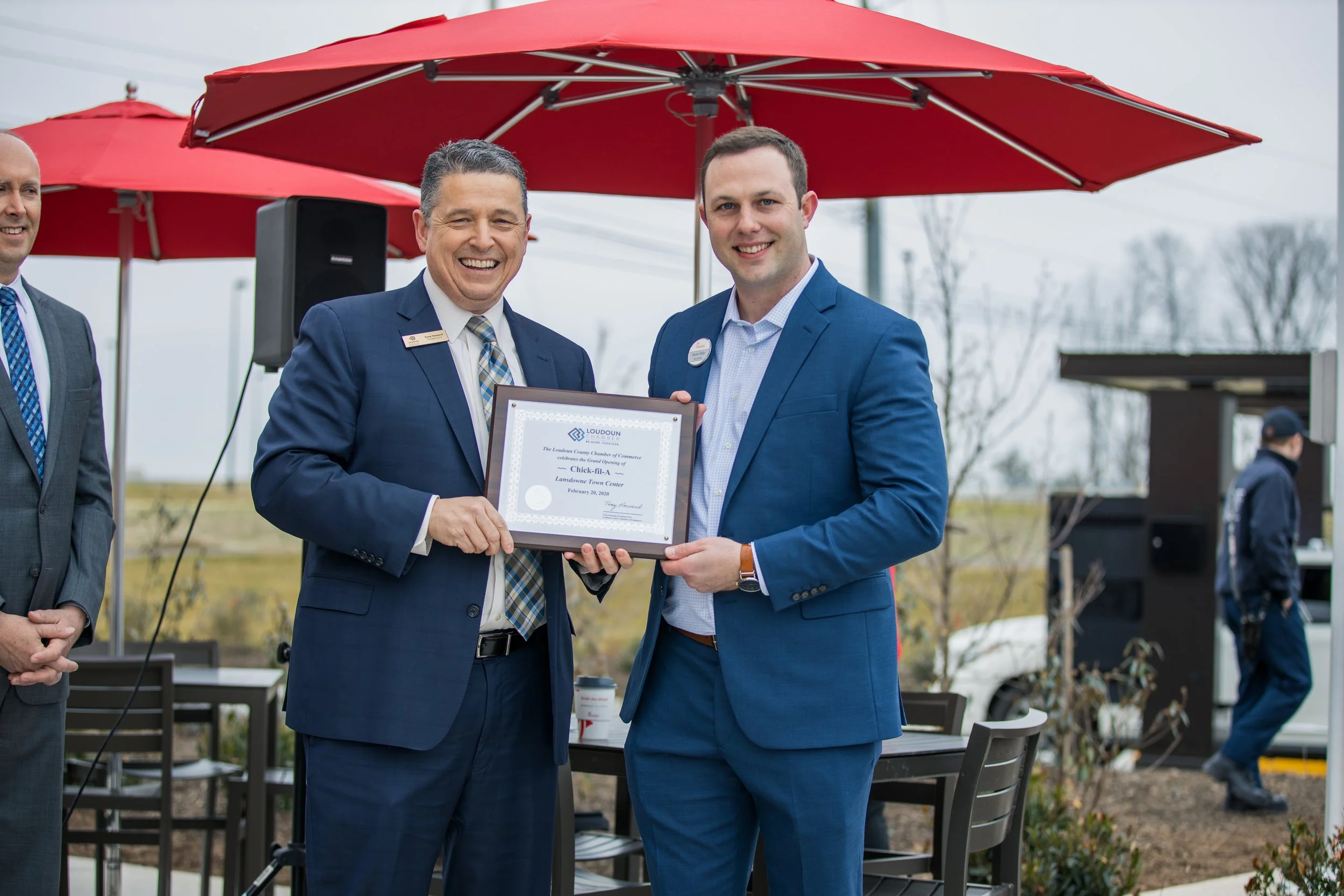 Two men in suits smiling and holding a framed certificate during an outdoor event, with red umbrellas and other people in the background. Grand opening in Ashburn Va. Event photography