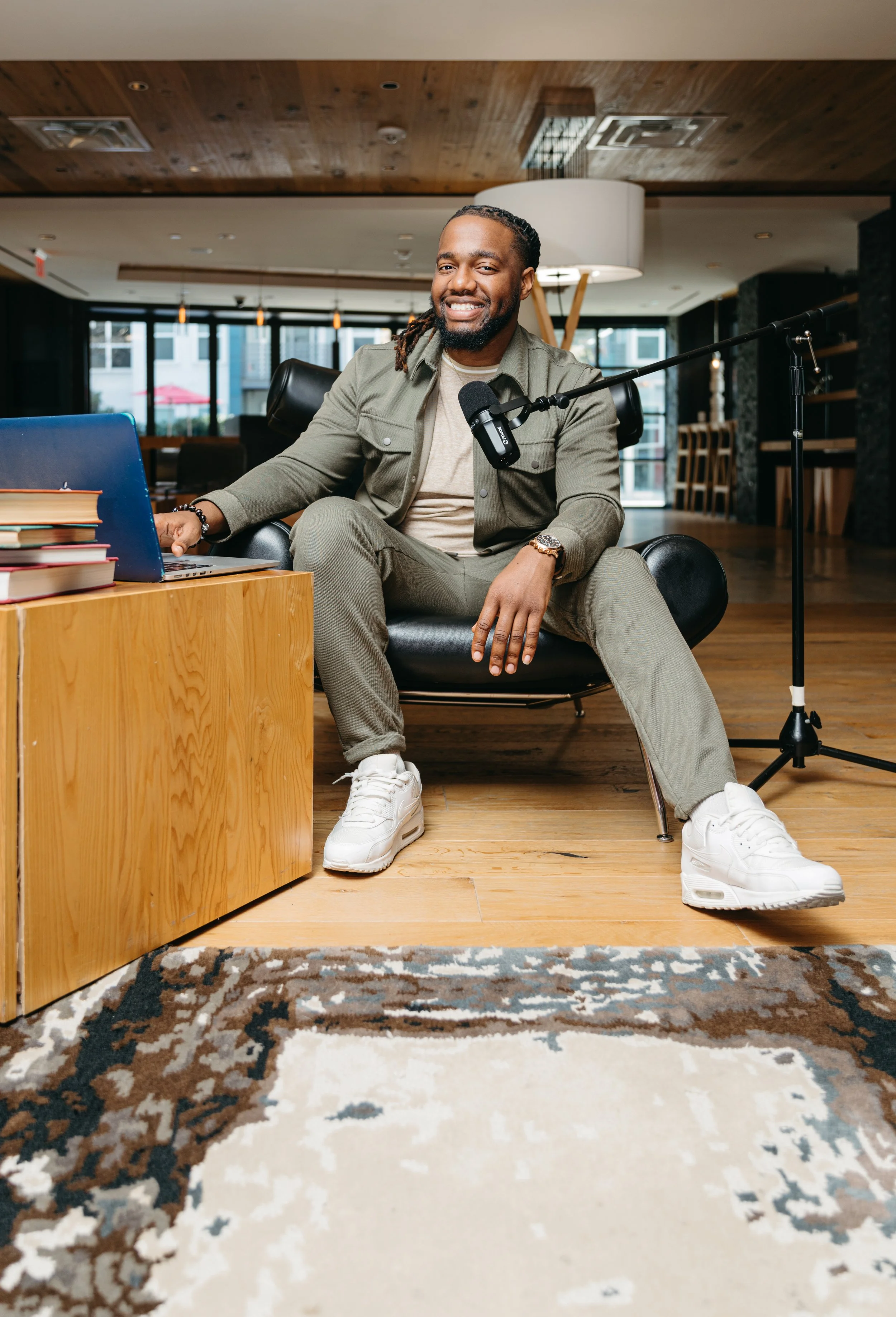 A man sitting on a black chair in a modern, well-lit indoor space, smiling, with a laptop on a wooden table, a microphone in front of him, and books on the table. Podcast Portraiture. Brand Portraits