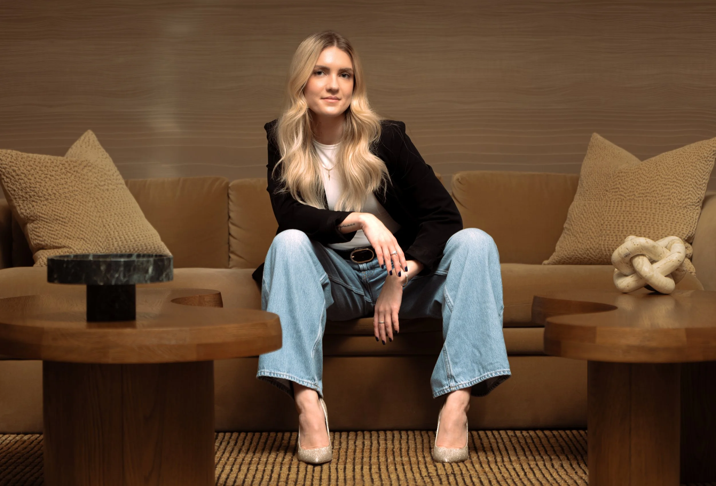 A woman with long blonde hair sitting on a tan sofa in a modern living room, wearing a black blazer, light blue jeans, and glittery heels, with a decorative knot sculpture and two textured pillows in the background. Luxury Hotel in Washington D.C. 