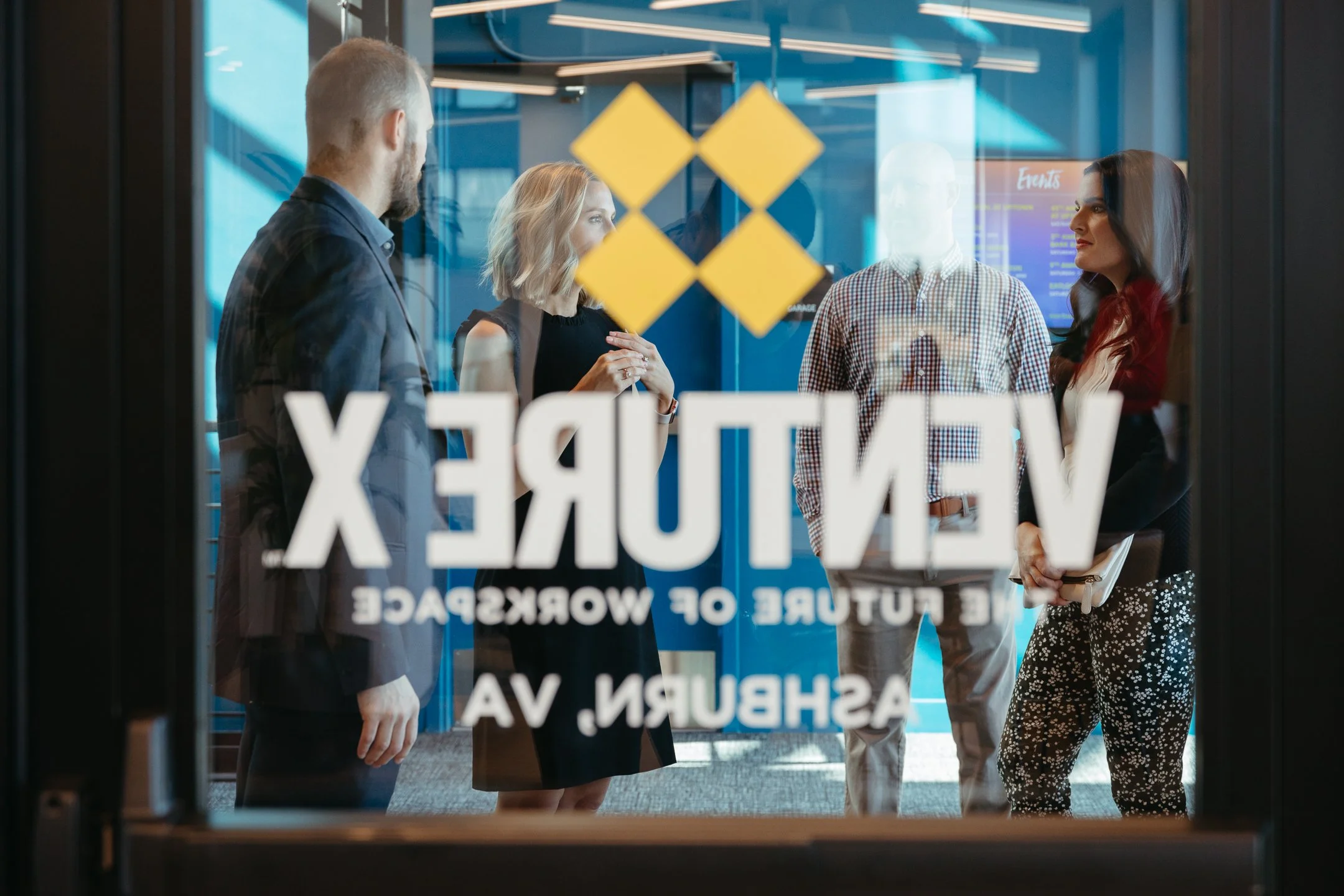 Group of people having a discussion in a modern office conference room, seen through frosted glass door with 'VentureX' and 'Founded of Workspace Ashley, VA' written on it.