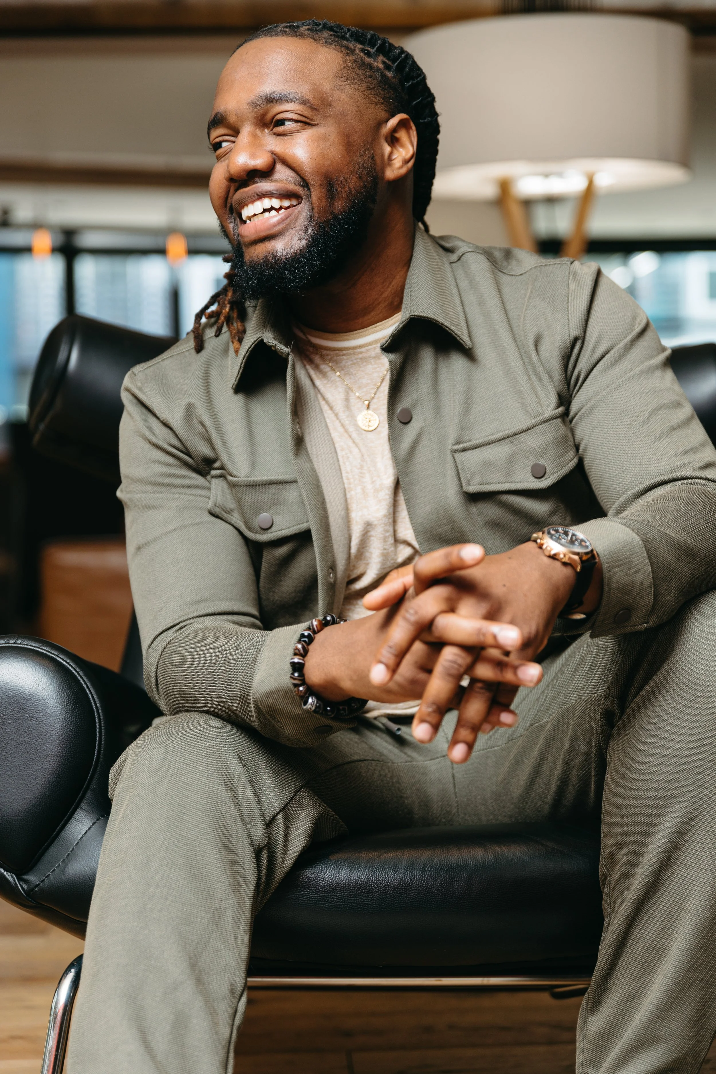 A smiling man sitting on a black chair in an indoor setting, wearing a green jacket, beige shirt, watch, and bracelet, with a blurred background of modern interior and windows. Brand Portraits
