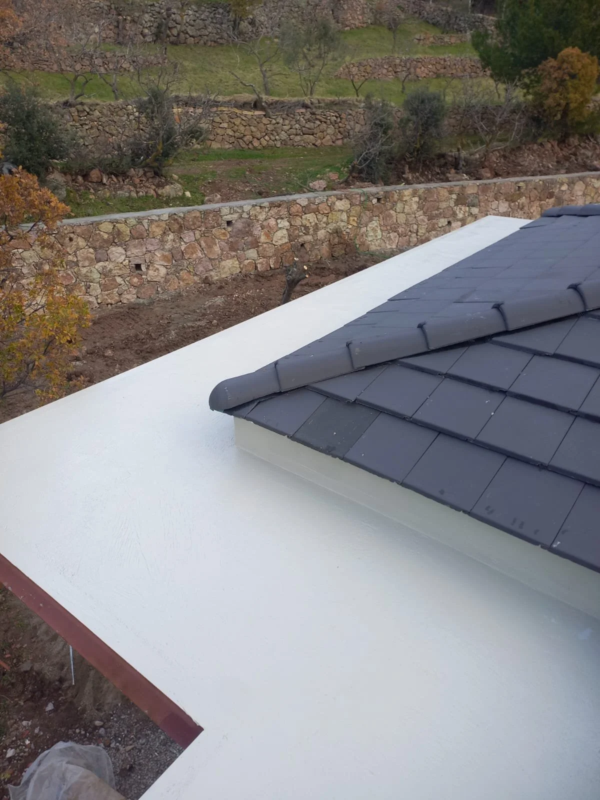 Beyaz bir çatı ve üst kısmında siyah kiremitli küçük bir yapı görülüyor, arka planda teraslar ve taş duvarlarla çevrili bahçe alanı var.