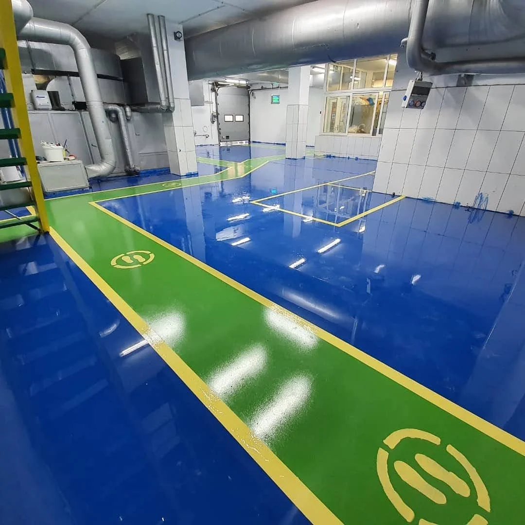 Fabrikaya ait temiz ve parlak mavi ve yeşil renkli zeminler, güvenlik alanlarını ve telefon simgelerini gösteren çizgiler ile işaretlenmiş iç mekan. Üst kısmında büyük hissettirici endüstriyel borular ve iki katlı cam duvarlar görülebilir.