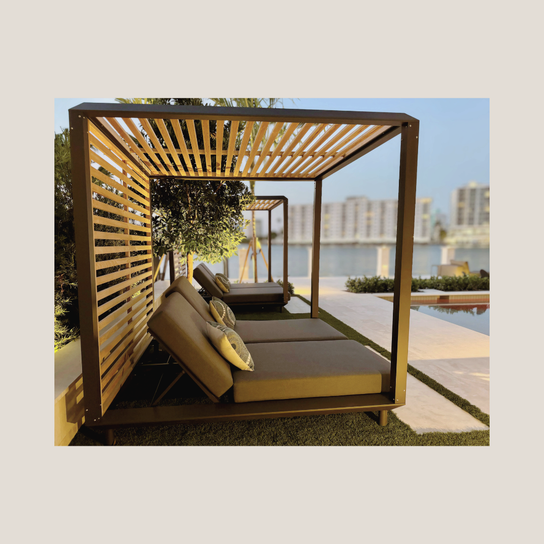 Outdoor seating area with two cushioned lounge beds under wooden pergolas near a pool, with a cityscape and water in the background.