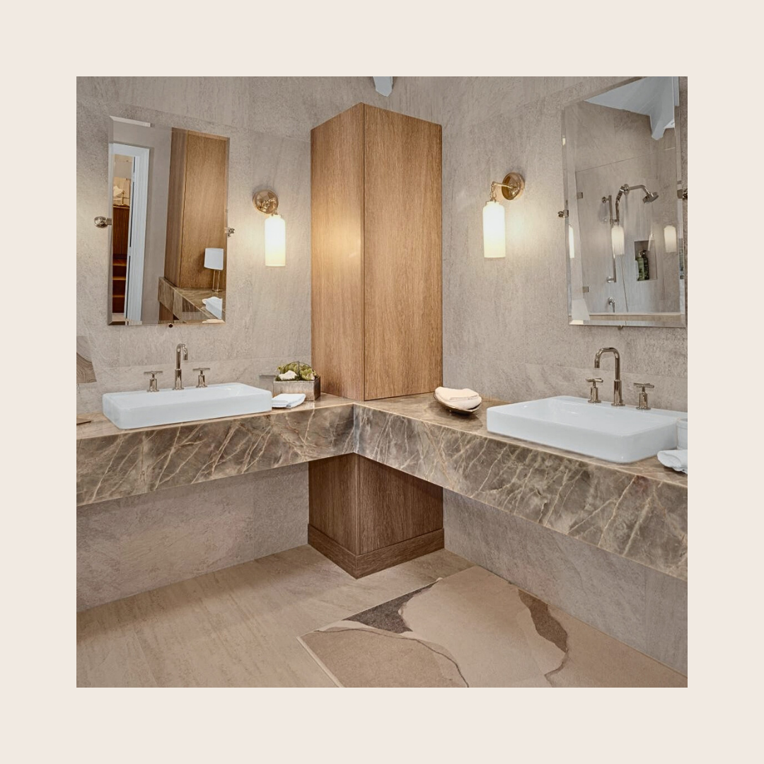 A modern bathroom with dual sinks, large mirrors, beige tiled walls, and a marble countertop. There are wall-mounted lights above each mirror and a walk-in shower with glass door in the background.