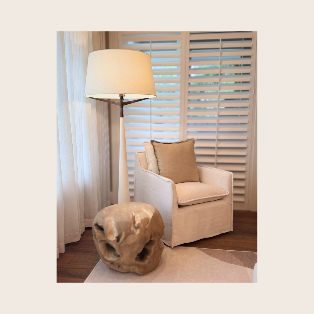 A cozy corner of a living room with a beige armchair, two pillows, a wooden side table, a tall floor lamp, beige curtains, and white plantation shutters on the window.