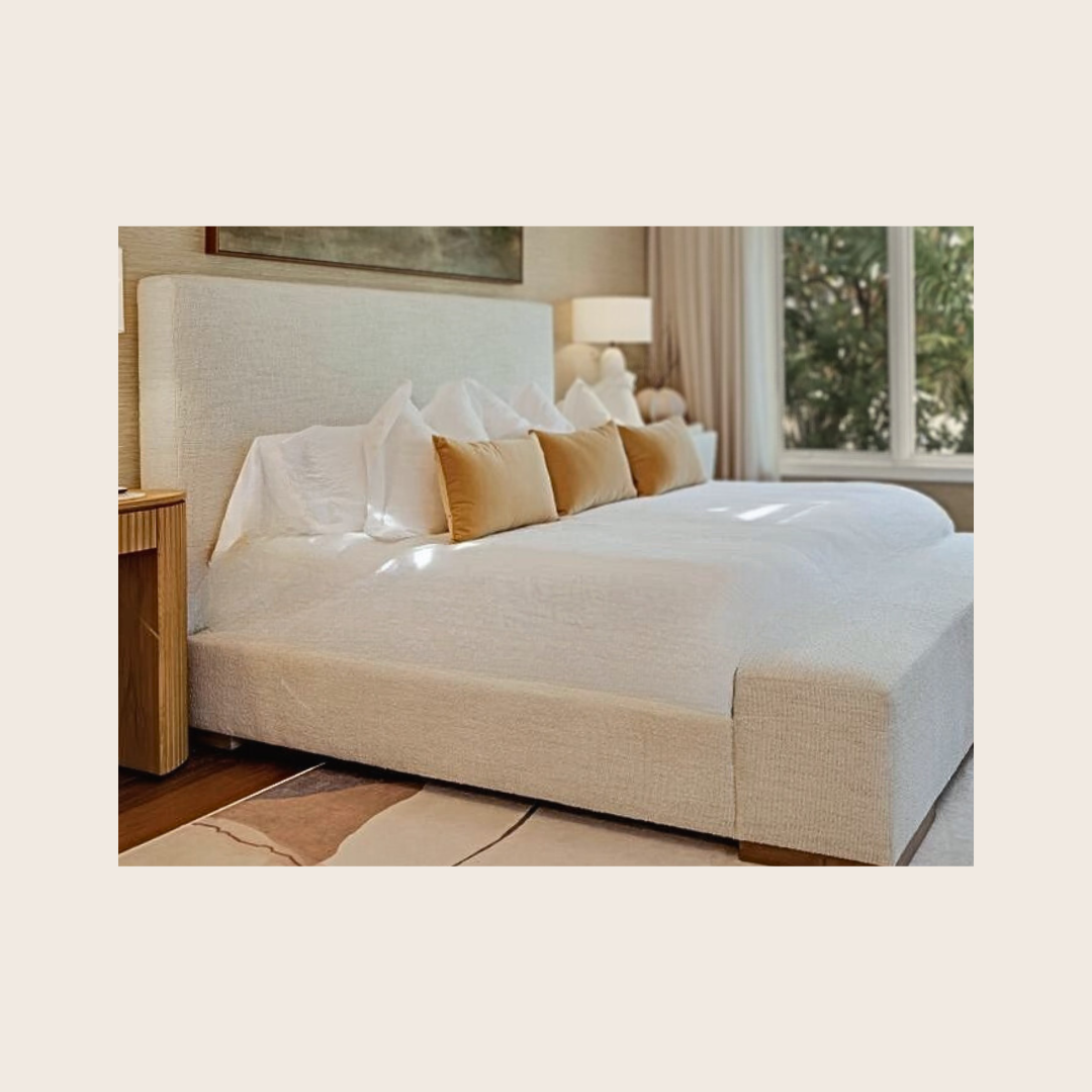 A neatly made bed with white bedding, multiple white pillows, and tan decorative pillows in a well-lit bedroom with a window showing trees outside.