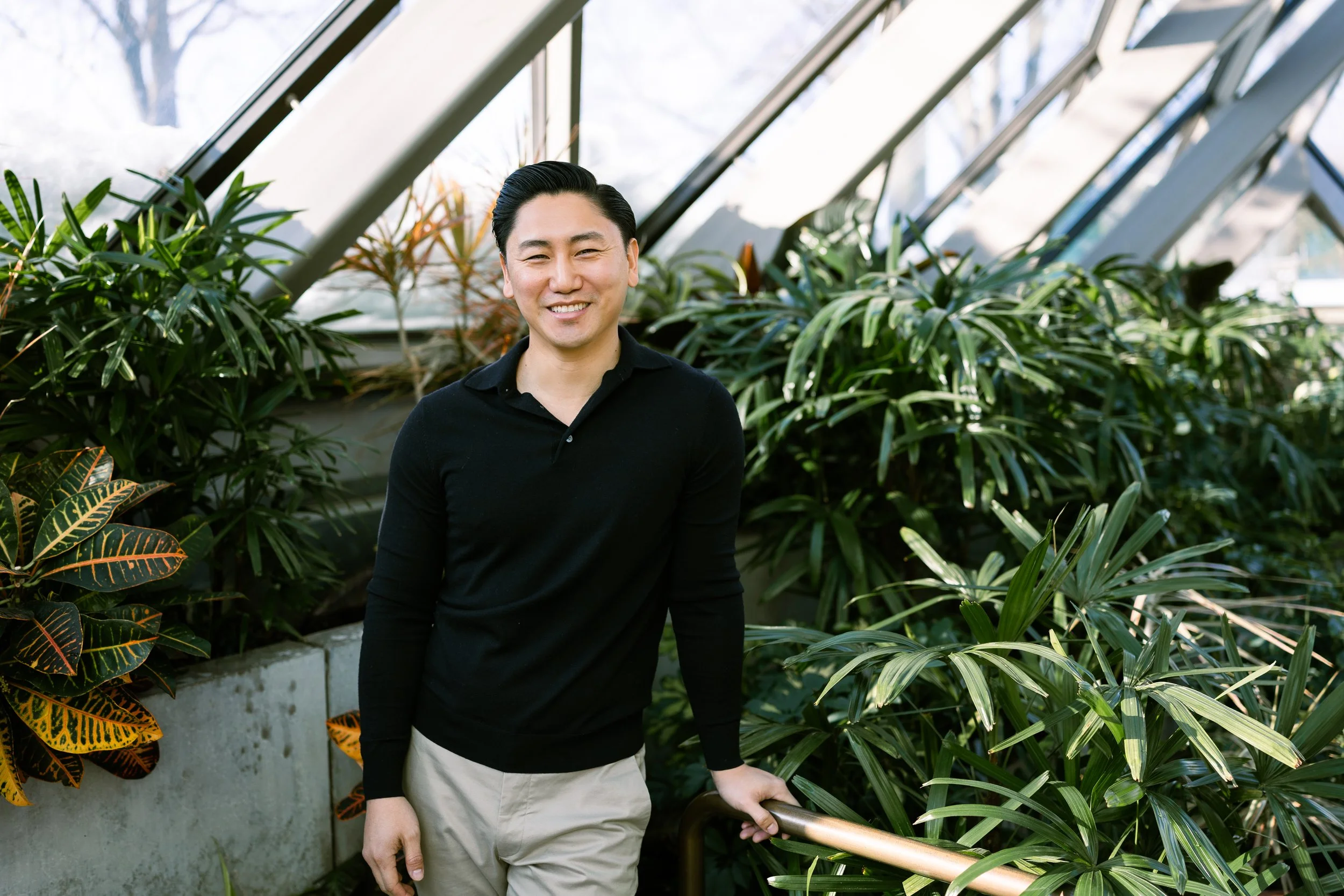A smiling man standing in a glass greenhouse surrounded by green and patterned tropical plants, wearing a black long-sleeve shirt and beige pants, holding a metal railing with one hand.