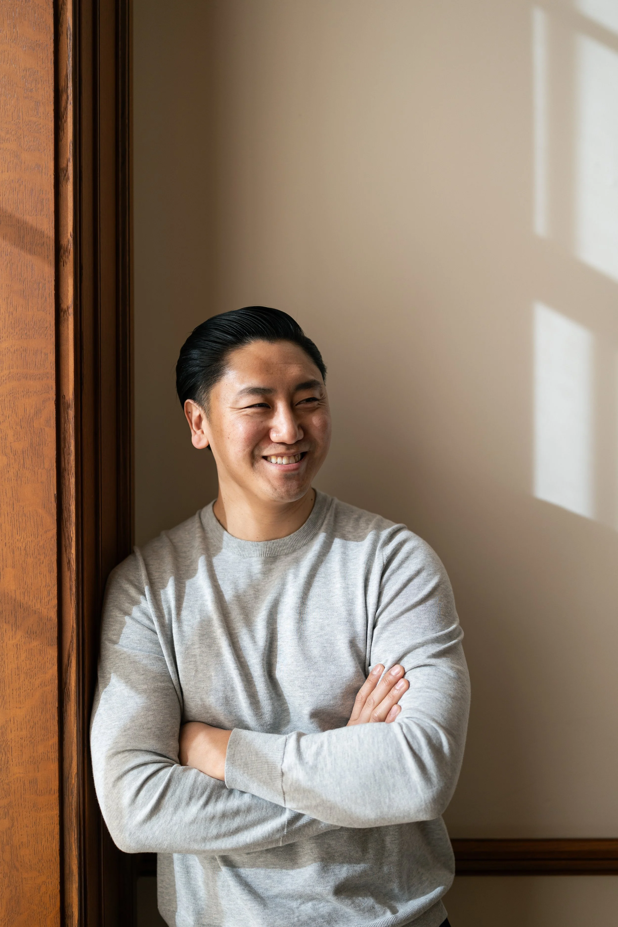 A young man with dark hair, smiling, standing with arms crossed beside a wooden door frame, with sunlight casting shadows from a window on the wall.