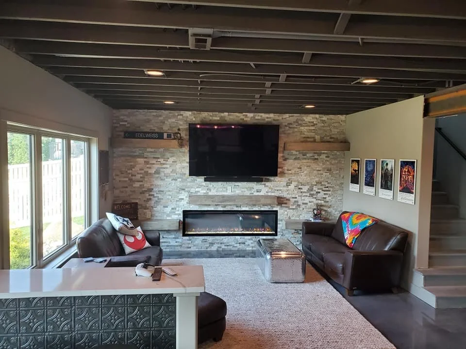 Living room with a stone accent wall, a large flat-screen TV above a modern fireplace, two leather sofas with colorful blankets, a small silver coffee table, and framed artwork on the walls.
