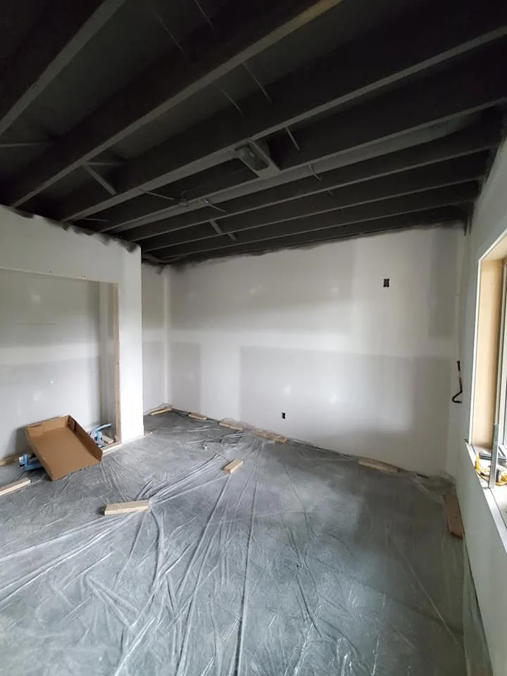 An unfinished interior room with drywall on the walls, plastic sheeting on the floor, and exposed ceiling beams under construction.