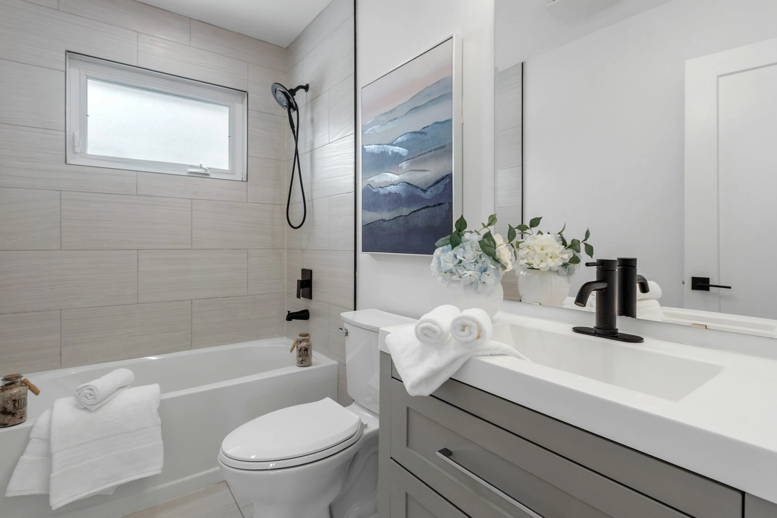Clean modern bathroom with bathtub, toilet, and vanity. Features include a small frosted window, floating black fixtures, a large mirror, and decorative white flowers with rolled towels.