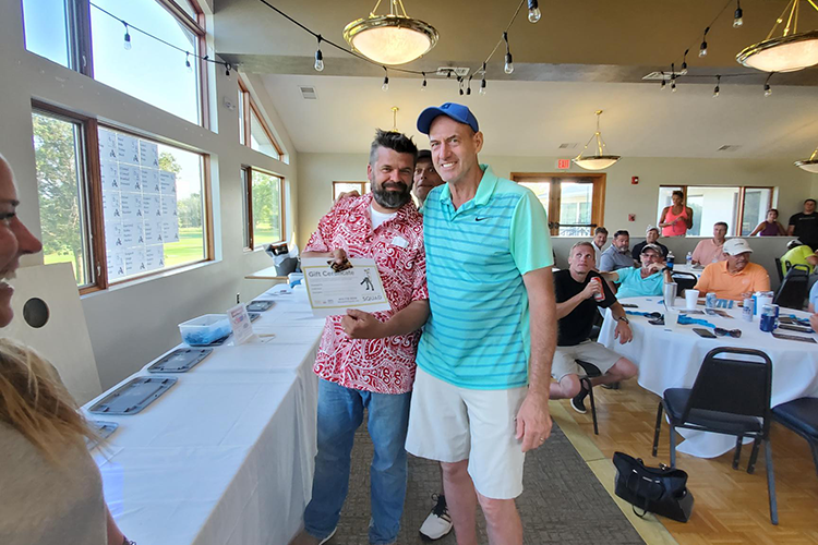 A man in a red patterned shirt holding a gift certificate and smiling next to a man in a teal striped polo shirt at an indoor event with seated guests.