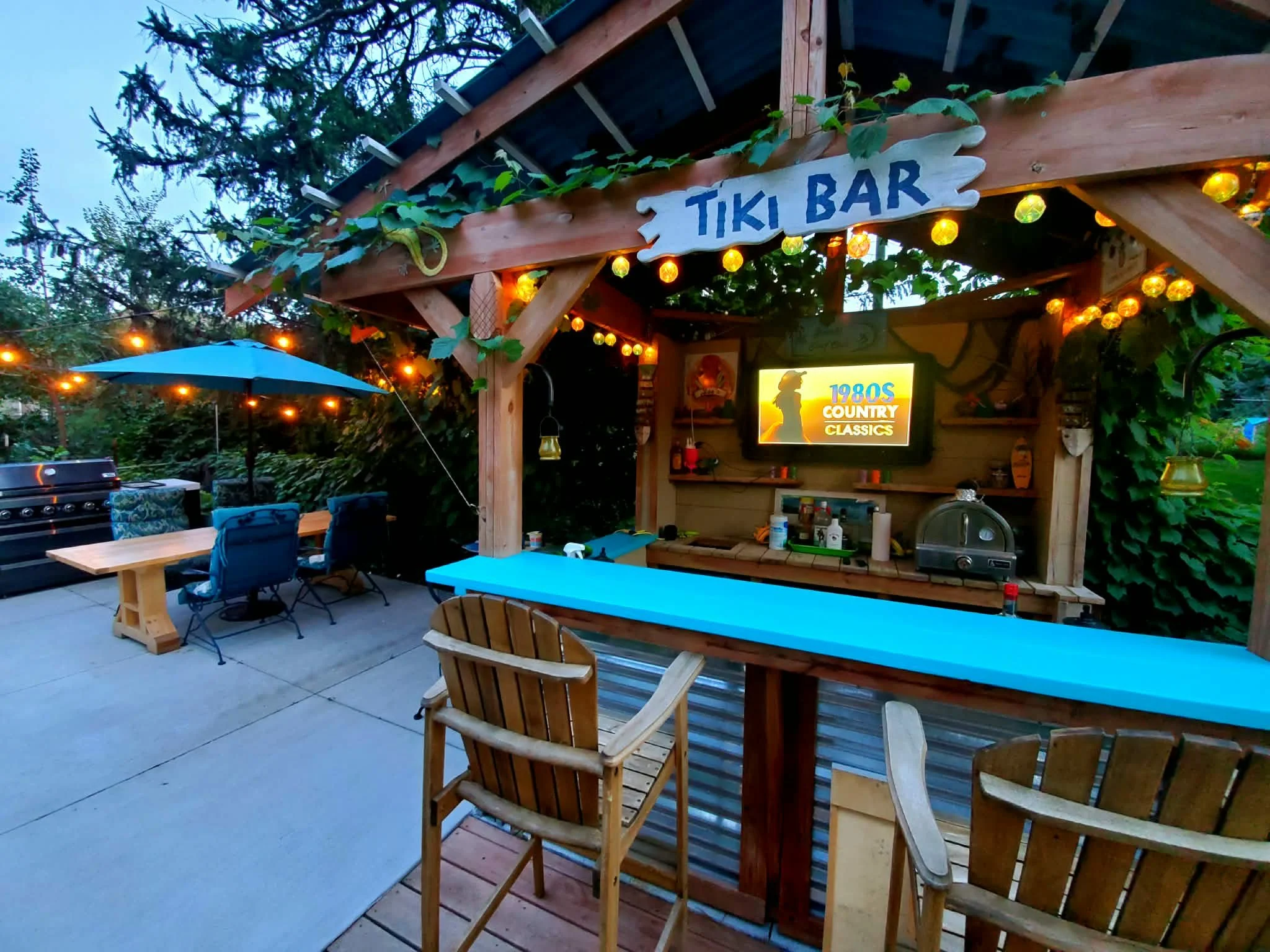Outdoor tiki bar with string lights, a blue umbrella, wooden chairs, a grill, and a television displaying '1980s country classics' surrounded by trees.