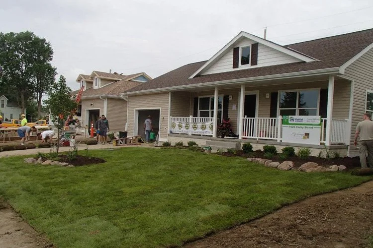 House with front porch, people working on landscaping, freshly planted grass, and construction activity.