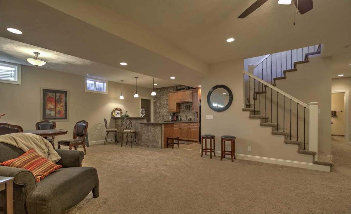 Living room with carpeted floor, staircase with carpeted steps, kitchen area with bar stools, and dining table with chairs, ceiling fan, and framed artwork on the wall.