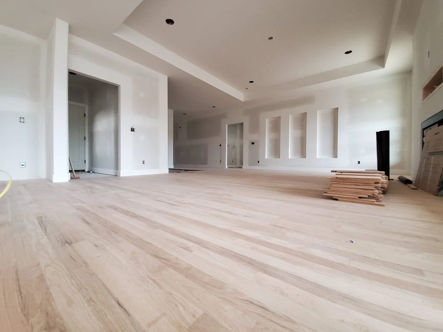 Interior of a room under construction with unfinished white walls and a light wooden floor, construction materials and tools scattered around.