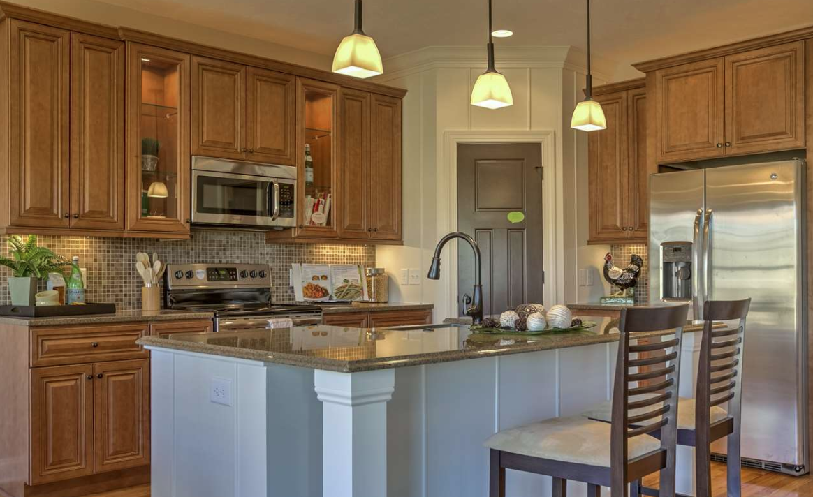 Kitchen with wooden cabinets, a stainless steel refrigerator, microwave, electric stove, granite countertops, island with a sink, pendant lights, and barstool chairs.