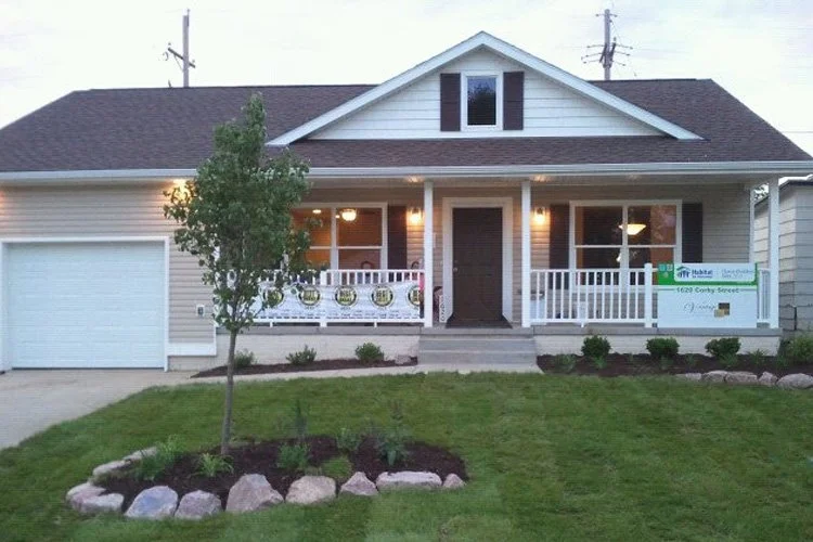 Front view of a house with a porch, white railing, brown front door, garage to the left, small tree and landscaped front yard.