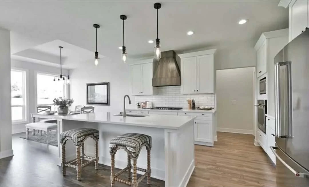 Modern white kitchen with an island, three pendant lights, stainless steel appliances, and a dining area with large windows.