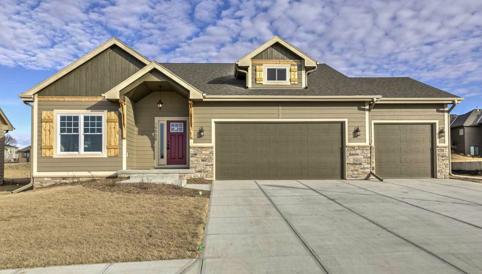 Front view of a modern house with a multi-gable roof, beige siding with stone accents, two-car garage, and a red front door. The house has a lawn and concrete driveway with a partly cloudy sky in the background.