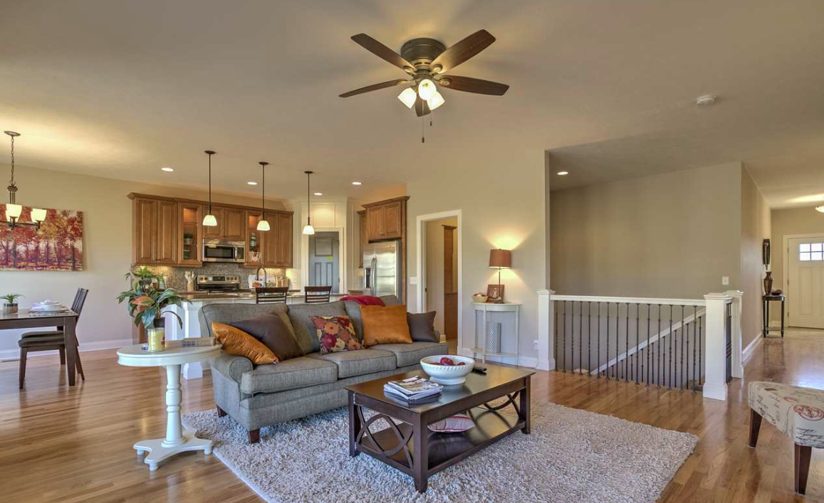 Open-concept living room and kitchen with hardwood floors, grey couch with decorative pillows, coffee table, dining table, pendant lights, ceiling fan, staircase, and front door.