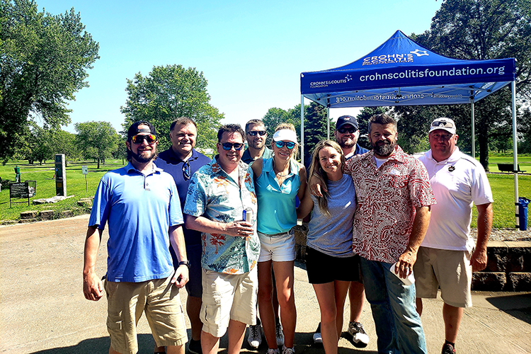 Group of nine people posing outdoors near a blue tent with 'Crohn's and Colitis Foundation' logo, on a sunny day in a park with trees.