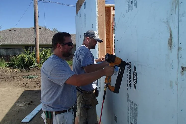 Two construction workers are installing insulation on a house wall with a nail gun. One worker is holding the nail gun while the other assists. They are outdoors on a building site.
