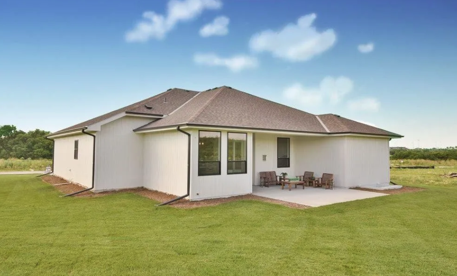 A white house with a brown roof, large front windows, and a small backyard patio with outdoor furniture, surrounded by green grass and open sky.