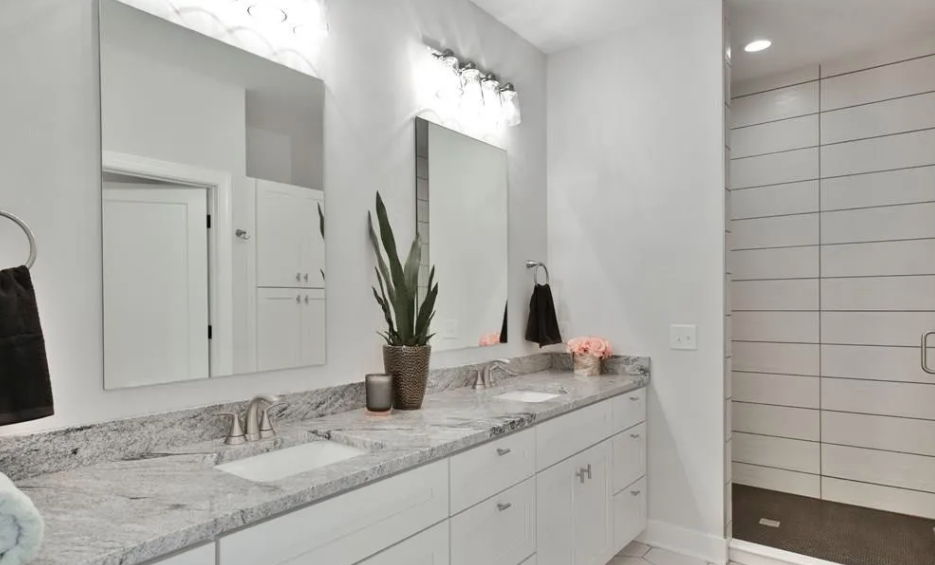 Modern bathroom with a white vanity, gray marble countertop, large mirror, and botanical decor. The shower has horizontal white tiles and a vestibule with white walls.