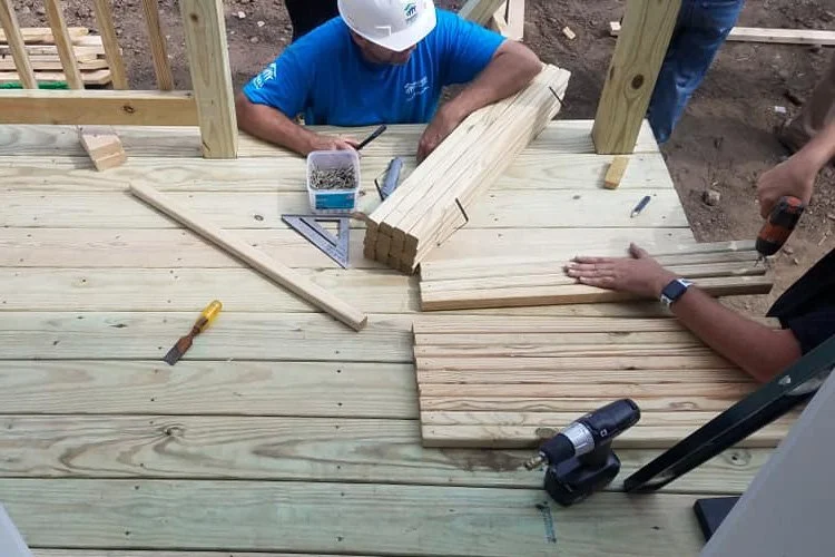 Workers building a wooden deck or floor, with tools and lumber on the surface.