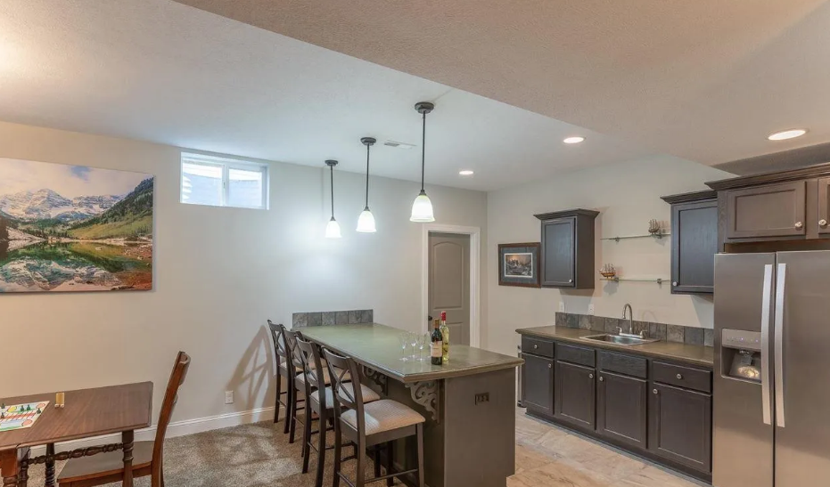 Kitchen with dark cabinets, a small island with three chairs, a stainless steel refrigerator, a sink, and a window. A dining area with a wooden table and chairs is partially visible, and there is a scenic landscape painting on the wall.
