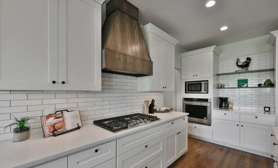 Kitchen with white cabinets, a gas stove, a wooden range hood, white subway tile backsplash, built-in microwave and oven, open shelves with decorative items, a potted plant, and wooden flooring.