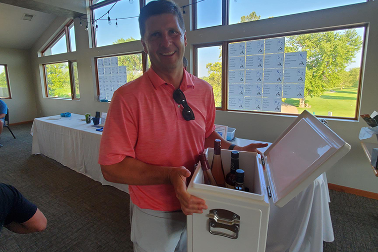 Man in pink polo shirt holding a cooler with wine bottles, standing in a room with large windows and a golf course view.