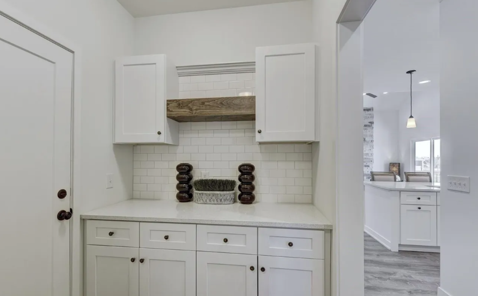 White kitchen with cabinets, a countertop, and decorative vases, visible through an open doorway.