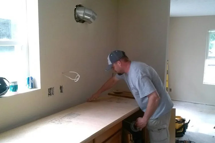 Man in a gray t-shirt and hat installing a wooden countertop in a room under construction, with electrical outlets and windows in the background.
