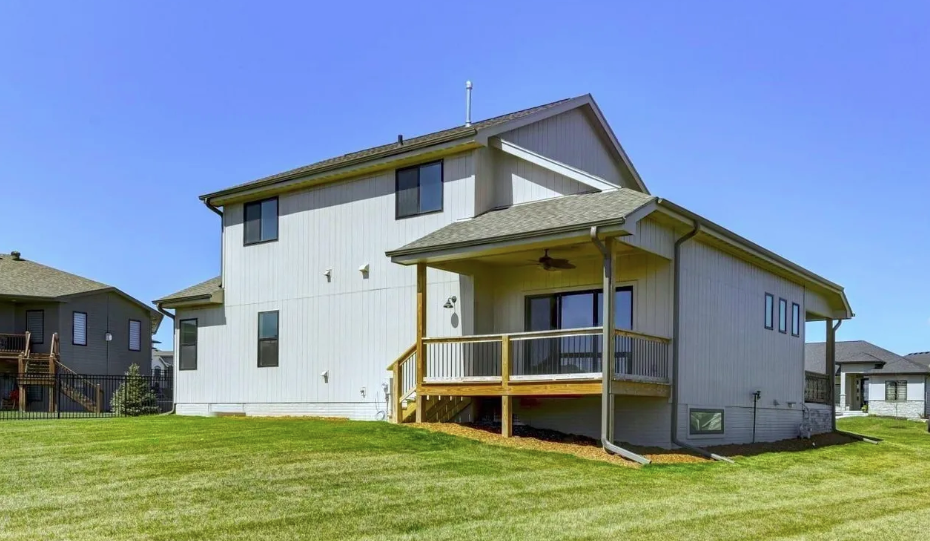 Backyard view of a modern two-story house with a small wooden deck, surrounded by green grass and neighboring houses, under a clear blue sky.