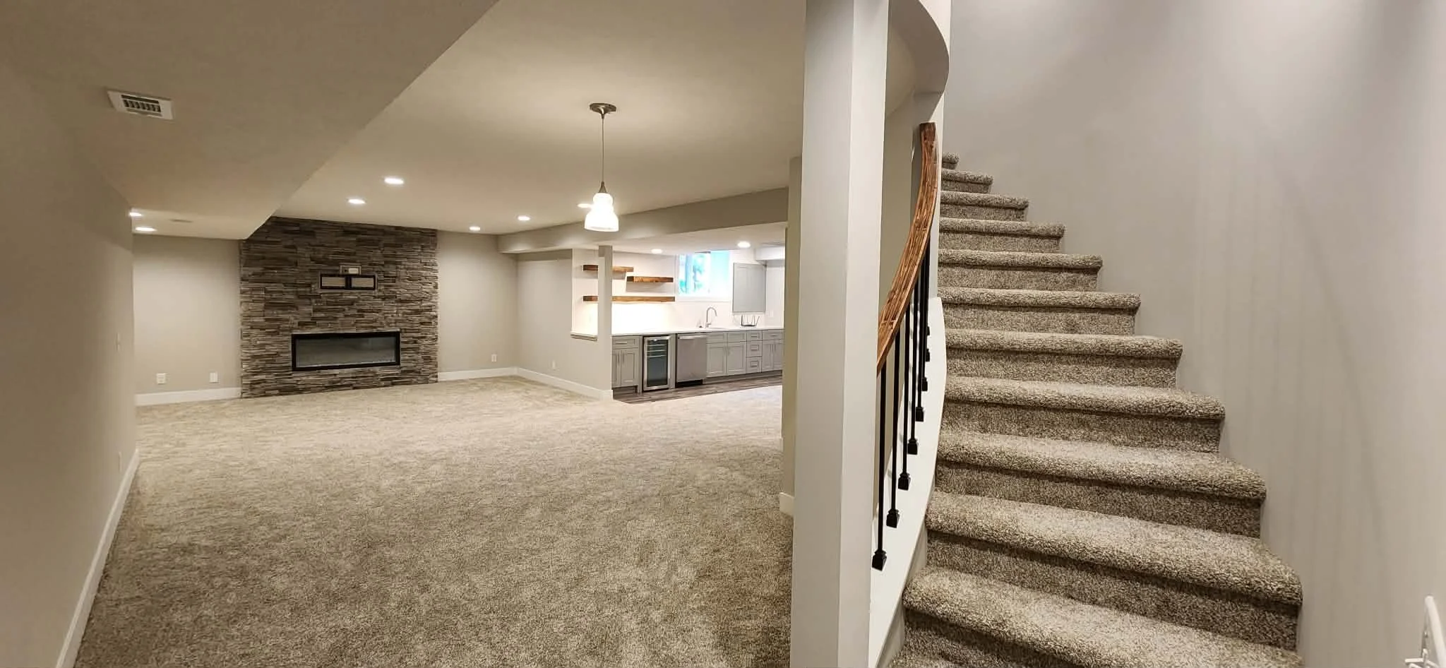 A spacious basement area with beige carpeting, a stone fireplace on the back wall, and a kitchenette with light gray cabinets, a small refrigerator, and a sink. There is a staircase to the right with carpeted steps and a wooden handrail. Recessed cei